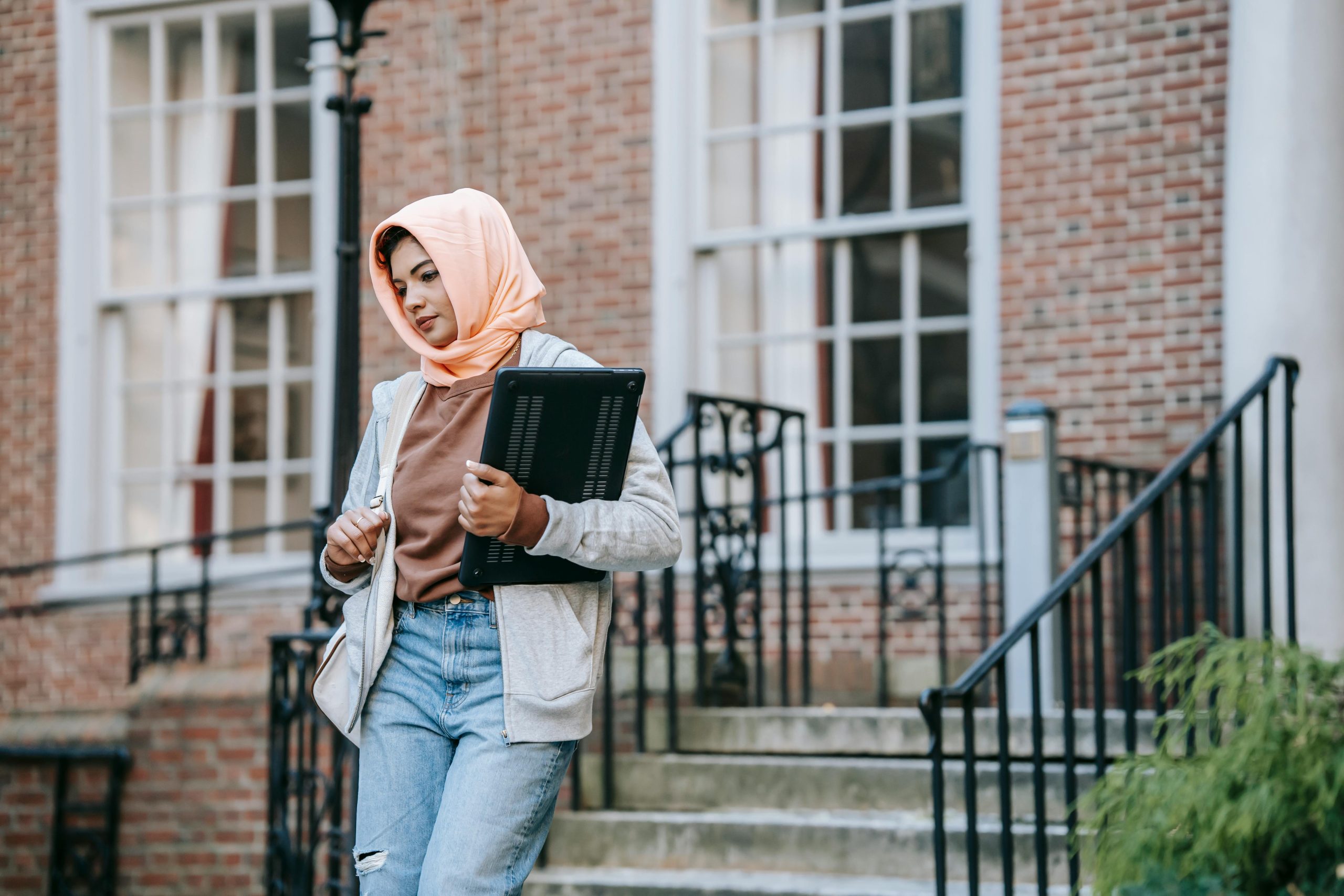 Young pensive Muslim female in casual wear and traditional headscarf and with laptop going down on stairs out of big building on street